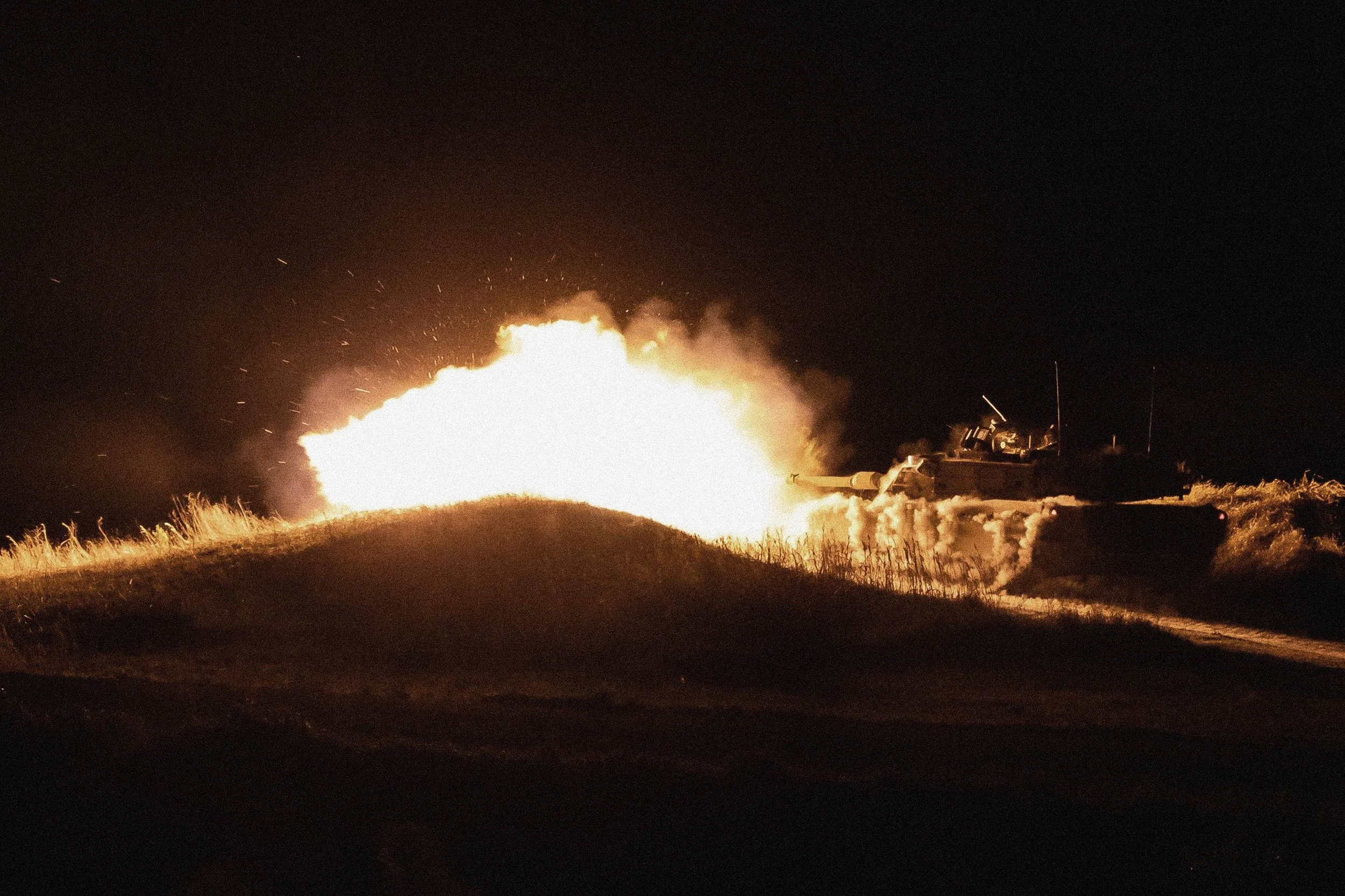 Nighttime scene of a military tank firing a large explosion on a grassy hill.