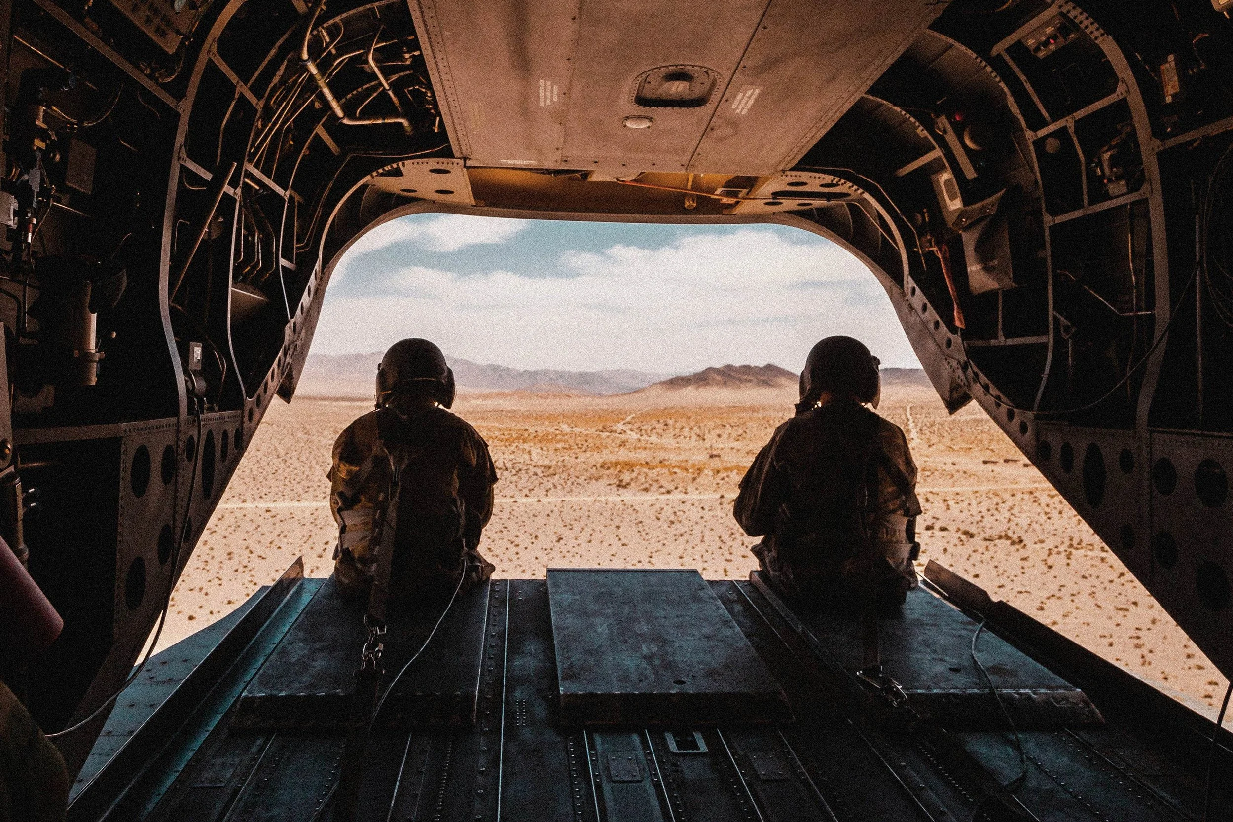 Two soldiers sitting in the open cargo door of a military helicopter flying over a desert landscape with mountains in the distance.